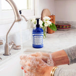 Hands washing with white soap lather over a sink, beside a blue Dr. Bronner's Peppermint Organic Sugar Soap 710ml pump bottle on a marble bench.