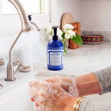 Hands washing with white soap lather over a sink, beside a blue Dr. Bronner's Peppermint Organic Sugar Soap 710ml pump bottle on a marble bench.
