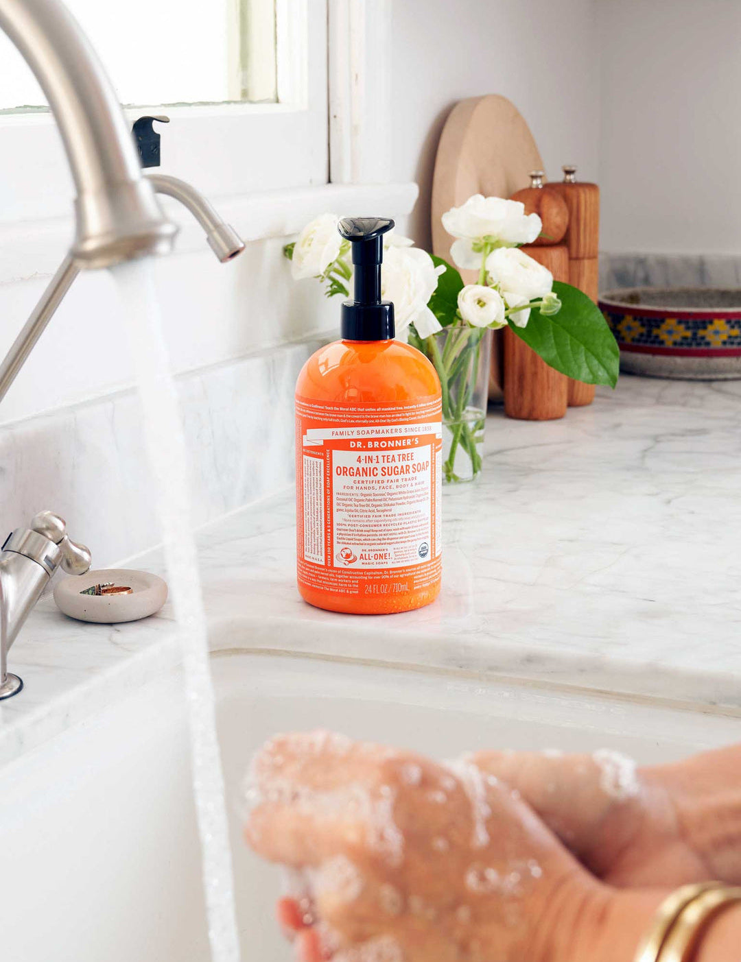 Hands rubbing together with soap lather over a white sink, beside an orange Dr. Bronner's Tea Tree Organic Sugar Soap 710ml pump bottle and white flowers.