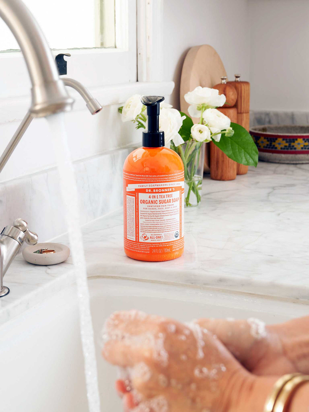 Hands rubbing together with soap lather over a white sink, beside an orange Dr. Bronner's Tea Tree Organic Sugar Soap 710ml pump bottle and white flowers.