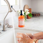 Hands rubbing together with soap lather over a white sink, beside an orange Dr. Bronner's Tea Tree Organic Sugar Soap 710ml pump bottle and white flowers.