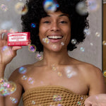 Smiling curly-haired woman wrapped in a brown towel holding a pink Dr. Bronner's Rose Pure-Castile Bar Soap packet among floating bubbles.