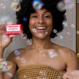 Smiling curly-haired woman wrapped in a brown towel holding a pink Dr. Bronner's Rose Pure-Castile Bar Soap packet among floating bubbles.