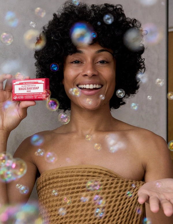 Smiling curly-haired woman wrapped in a brown towel holding a pink Dr. Bronner's Rose Pure-Castile Bar Soap packet among floating bubbles.