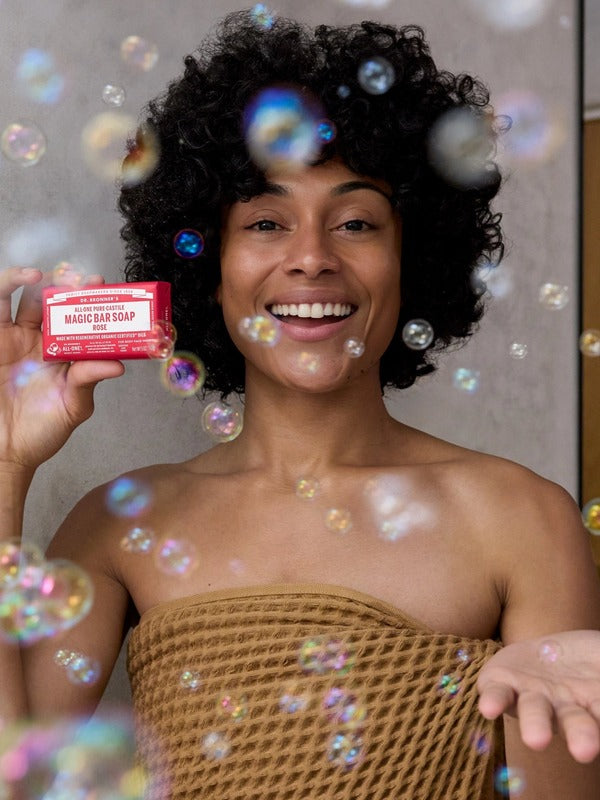 Smiling curly-haired woman wrapped in a brown towel holding a pink Dr. Bronner's Rose Pure-Castile Bar Soap packet among floating bubbles.