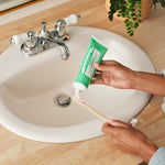 Hands squeezing green Dr. Bronner's Spearmint Toothpaste onto a wooden toothbrush above a white bathroom basin.