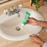 Hands squeezing green Dr. Bronner's Spearmint Toothpaste onto a wooden toothbrush above a white bathroom basin.