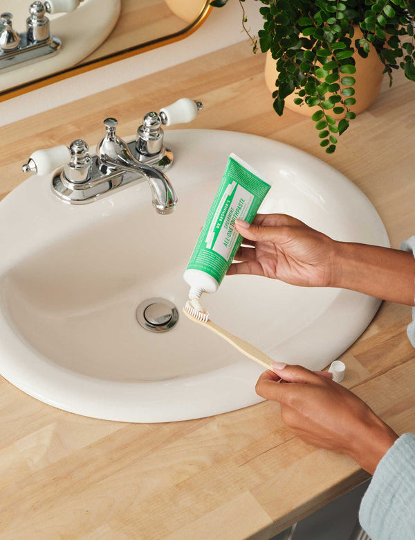 Hands squeezing green Dr. Bronner's Spearmint Toothpaste onto a wooden toothbrush above a white bathroom basin.