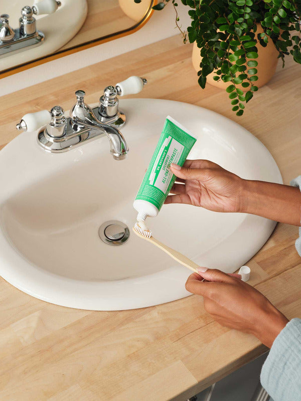 Hands squeezing green Dr. Bronner's Spearmint Toothpaste onto a wooden toothbrush above a white bathroom basin.