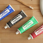 Four Dr. Bronner's All-One Toothpaste tubes arranged diagonally on a wooden counter: blue Peppermint, black Anise, green Spearmint and red Cinnamon, with a wooden toothbrush near a white basin.