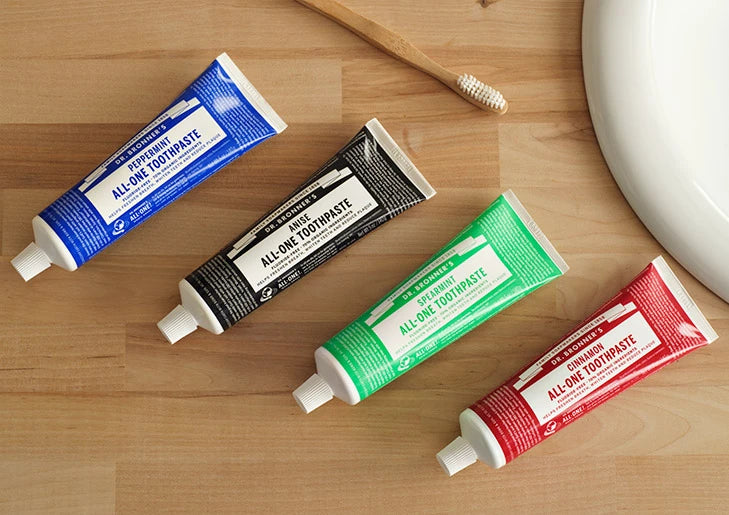 Four Dr. Bronner's All-One Toothpaste tubes arranged diagonally on a wooden counter: blue Peppermint, black Anise, green Spearmint and red Cinnamon, with a wooden toothbrush near a white basin.