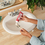 Hands squeezing toothpaste from a red Dr. Bronner's Cinnamon tube onto a wooden toothbrush above a white bathroom basin.