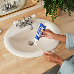Hands squeezing toothpaste from a blue Dr. Bronner's Peppermint tube onto a wooden toothbrush above a white bathroom basin.