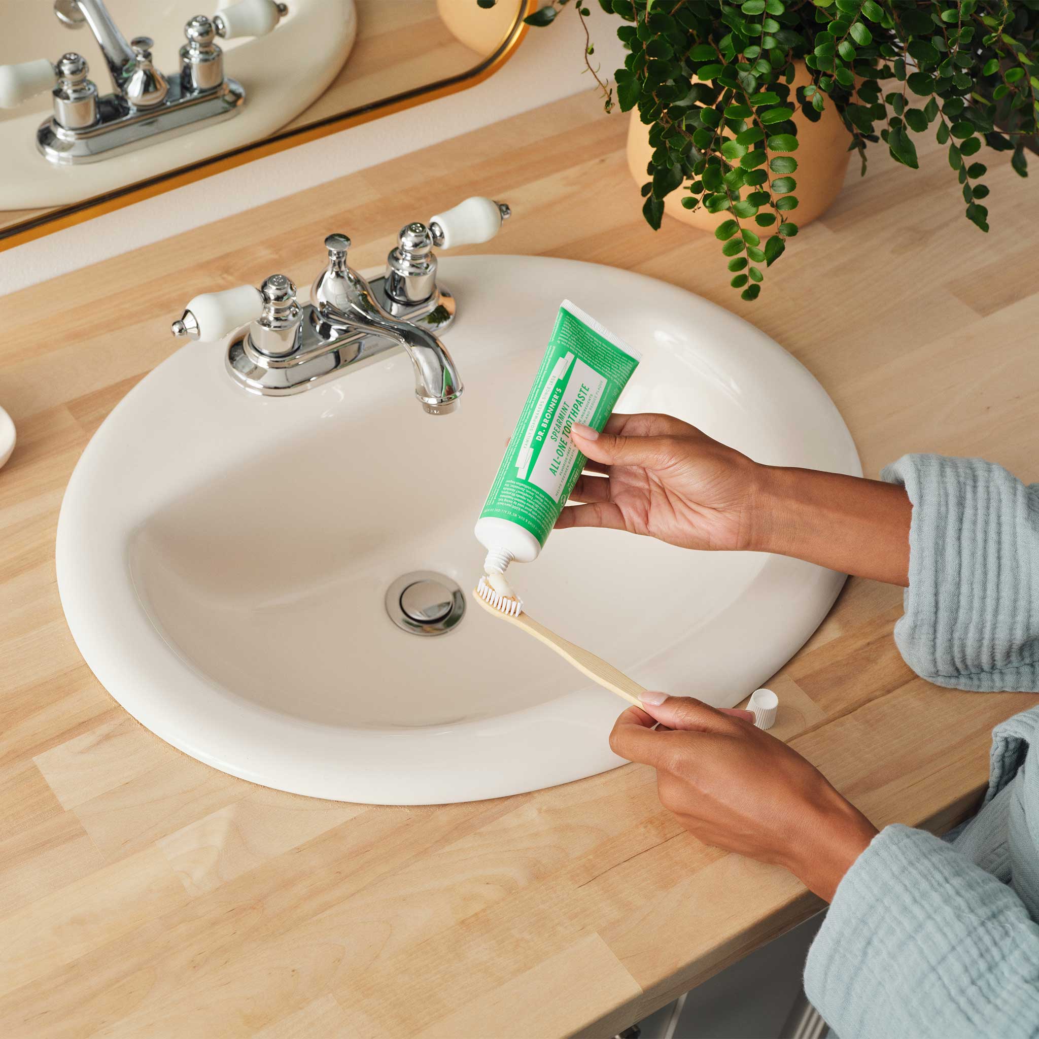 Hands squeezing toothpaste from a green Dr. Bronner's Spearmint tube onto a wooden toothbrush above a white bathroom basin.