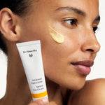 Woman holding a white Dr Hauschka Apricot Day Cream tube beside her face, with a glossy golden-yellow cream swatch applied to her cheek.