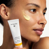 Woman holding a white Dr Hauschka Apricot Day Cream tube beside her face, with a glossy golden-yellow cream swatch applied to her cheek.