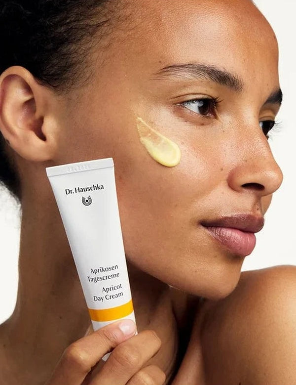 Woman holding a white Dr Hauschka Apricot Day Cream tube beside her face, with a glossy golden-yellow cream swatch applied to her cheek.