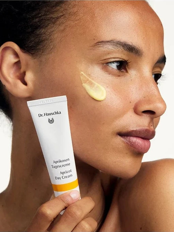 Woman holding a white Dr Hauschka Apricot Day Cream tube beside her face, with a glossy golden-yellow cream swatch applied to her cheek.