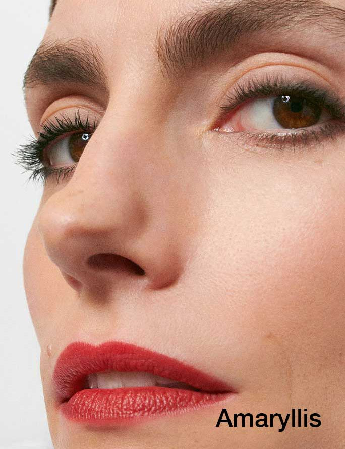 Close-up of a face with brown eyes, long lashes, defined brows, and red lips wearing Dr Hauschka Lipsticks 4g made with hydrating botanicals. "Amaryllis" is written in black text at the bottom right corner.