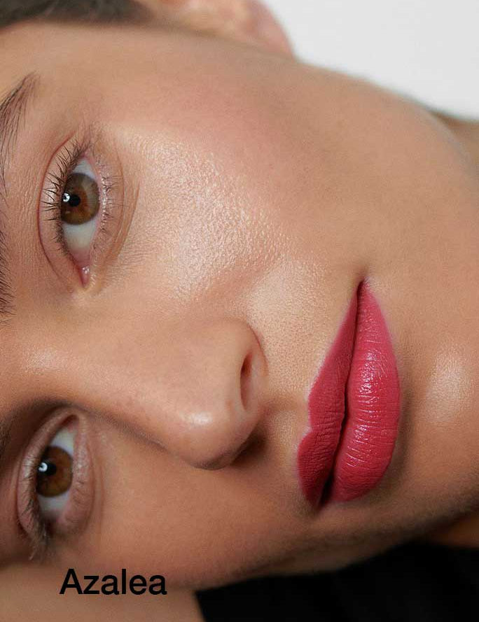 A close-up of a woman’s smooth-skinned face with natural makeup, wearing bold pink Dr Hauschka Lipsticks 4g made with hydrating botanical ingredients. "Azalea" appears in black text in the lower left corner.