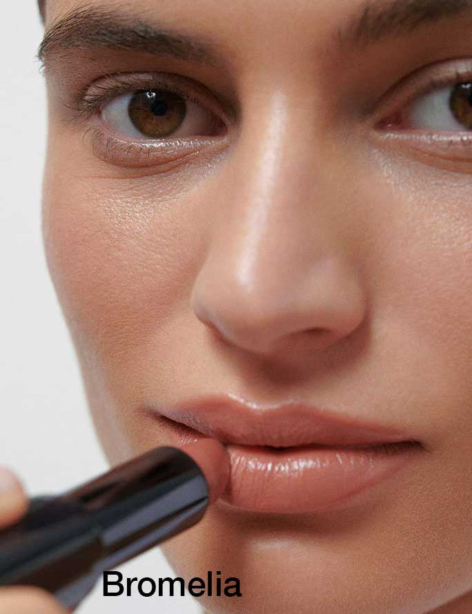 A close-up of a person applying Dr Hauschka Lipsticks 4g in a nude shade shows smooth skin and brown eyes. The hydrating formula features botanical ingredients. "Bromelia" is written in black text at the bottom left.