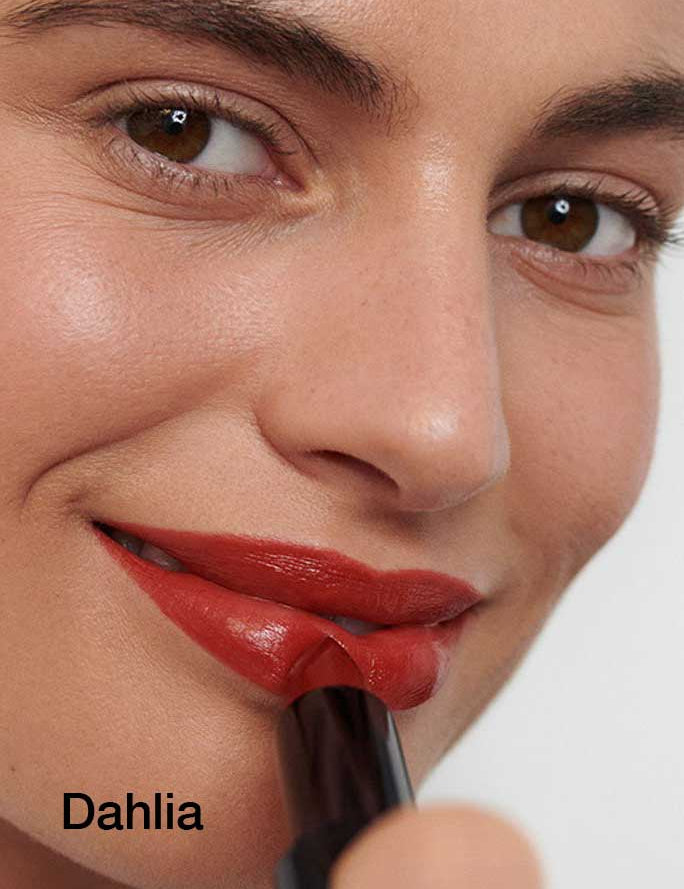 A person with smooth skin and brown eyes applies Dr Hauschka Lipsticks 4g in dark red, smiling slightly. The lipstick features hydrating botanical ingredients. "Dahlia" is written in black in the lower left corner.