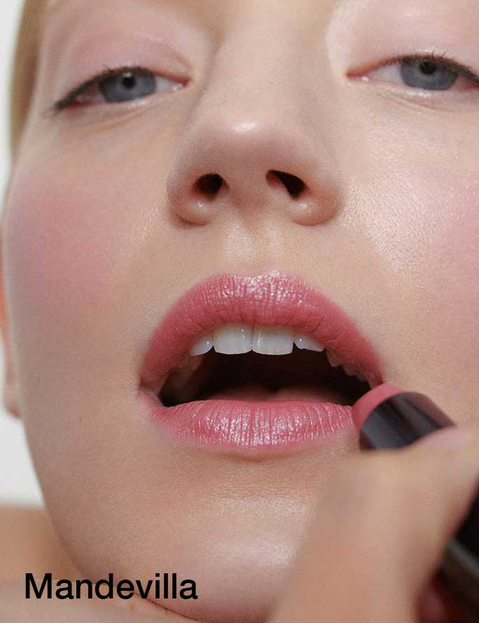 A close-up of a person applying Dr Hauschka Lipsticks 4g to slightly open lips, emphasizing hydrating botanical ingredients. “Mandevilla” is shown in black text at the bottom left corner.