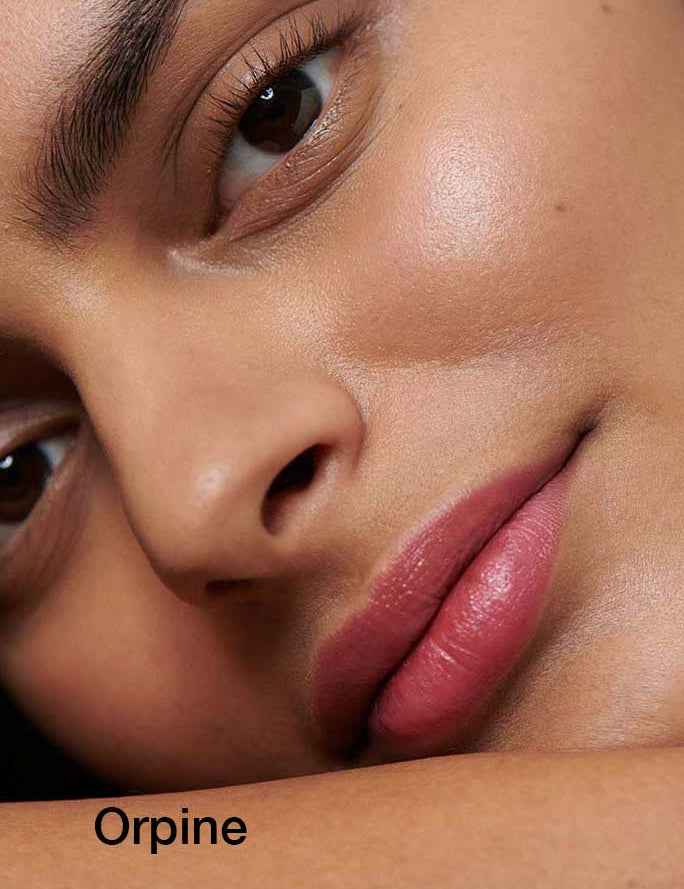 A close-up of a woman’s radiant skin and natural makeup, highlighting her eye, nose, and pink lips. “Dr Hauschka Lipsticks 4g” appears below, showcasing hydrating botanical ingredients for naturally beautiful lips.