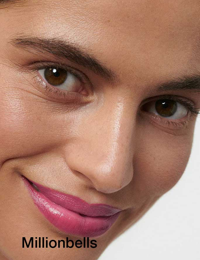 A close-up of a person with smooth skin, brown eyes, and softly smiling lips wearing Dr Hauschka Lipsticks 4g with hydrating botanical ingredients. The word Millionbells appears in black text at the bottom left.