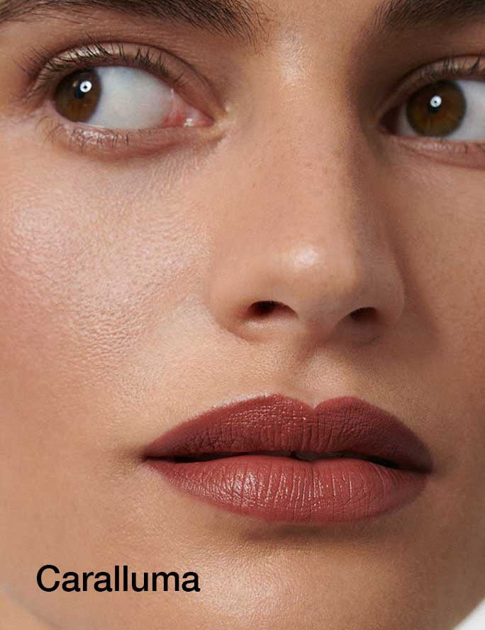 A close-up of a face with smooth skin, defined brows, brown eyes looking to the side, and matte rose lips achieved with Dr Hauschka Lipsticks 4g. The word Caralluma appears in black text at the bottom left.