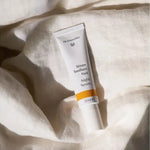 White Dr Hauschka Night Serum tube resting upright among softly folded white linen sheets in warm natural light.