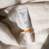 White Dr Hauschka Night Serum tube resting upright among softly folded white linen sheets in warm natural light.