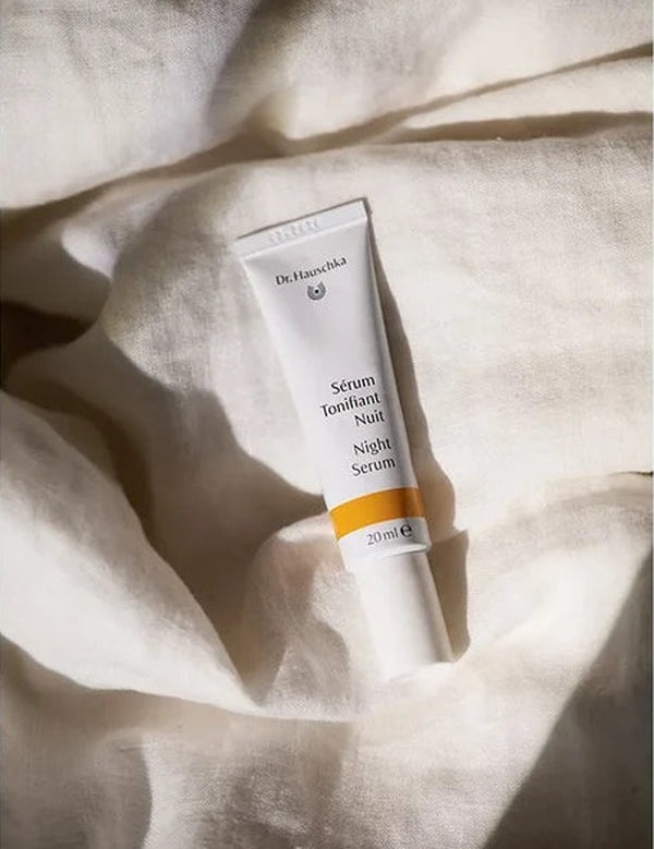White Dr Hauschka Night Serum tube resting upright among softly folded white linen sheets in warm natural light.