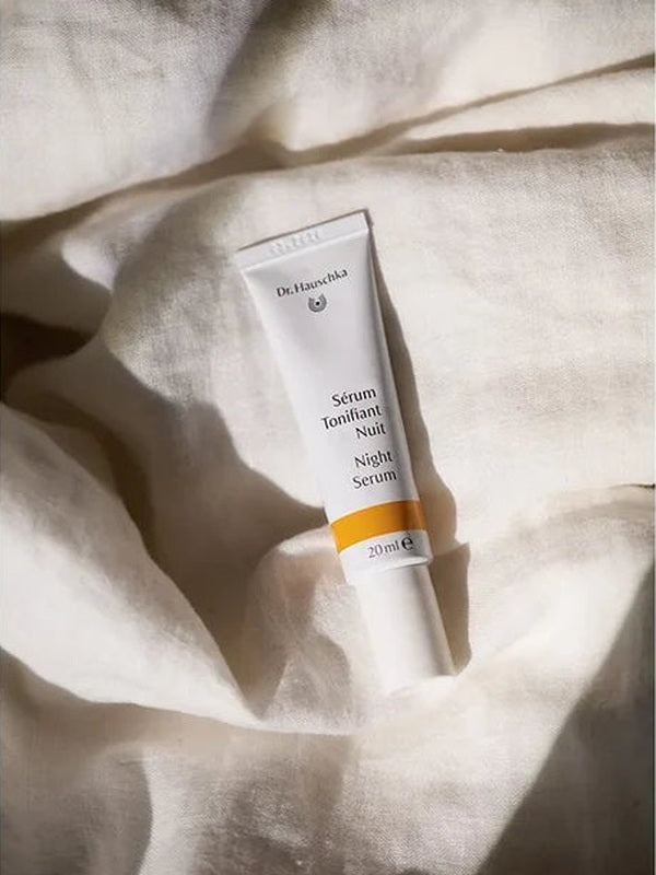 White Dr Hauschka Night Serum tube resting upright among softly folded white linen sheets in warm natural light.