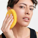 Woman pressing a golden-yellow Dr Hauschka Oval Cleansing Sponge against her cheek while cleansing her face.