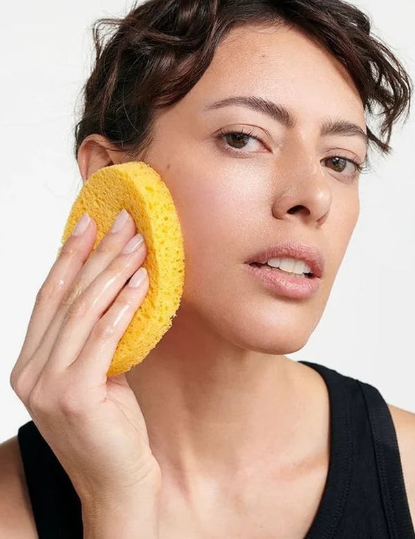 Woman pressing a golden-yellow Dr Hauschka Oval Cleansing Sponge against her cheek while cleansing her face.