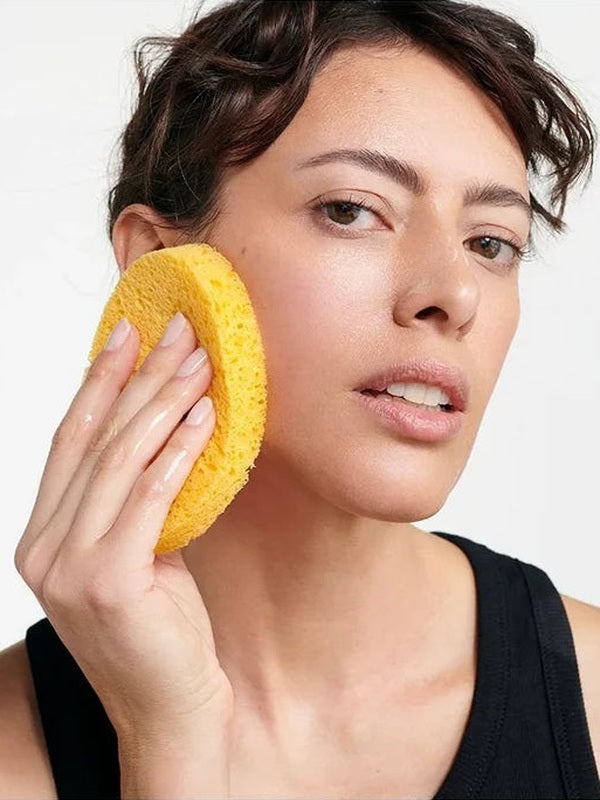 Woman pressing a golden-yellow Dr Hauschka Oval Cleansing Sponge against her cheek while cleansing her face.