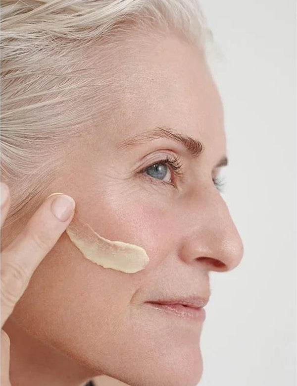 Grey-haired woman applying a thick ivory streak of Dr Hauschka Regenerating Day Cream Intensive 40ml across her cheek.