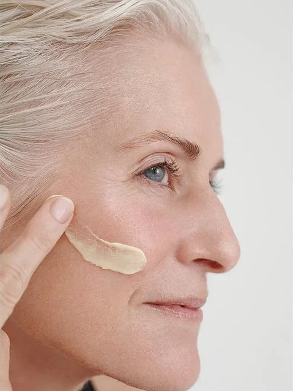 Grey-haired woman applying a thick ivory streak of Dr Hauschka Regenerating Day Cream Intensive 40ml across her cheek.