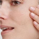 Close-up of a model smoothing a clear streak of Dr Hauschka Regenerating Serum 30ml across her cheek with two fingertips.