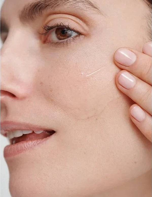 Close-up of a model smoothing a clear streak of Dr Hauschka Regenerating Serum 30ml across her cheek with two fingertips.
