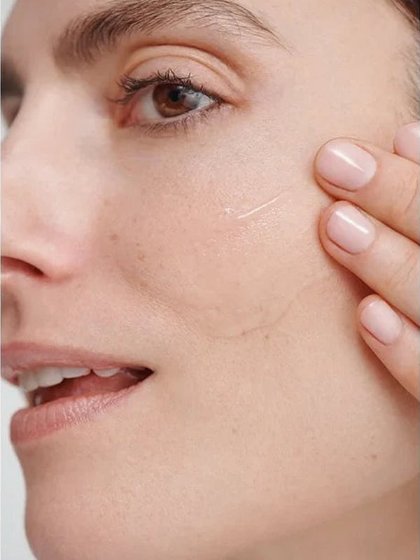 Close-up of a model smoothing a clear streak of Dr Hauschka Regenerating Serum 30ml across her cheek with two fingertips.