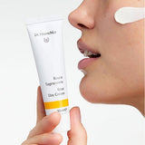 Woman holding a white Dr Hauschka Rose Day Cream tube beside her face, with an opaque white cream swatch on her cheek.