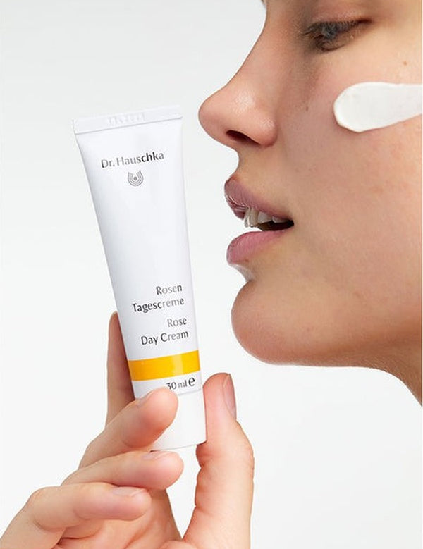 Woman holding a white Dr Hauschka Rose Day Cream tube beside her face, with an opaque white cream swatch on her cheek.