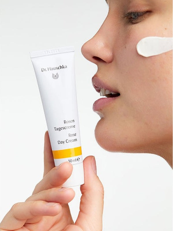 Woman holding a white Dr Hauschka Rose Day Cream tube beside her face, with an opaque white cream swatch on her cheek.