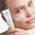 Woman holding a white Dr Hauschka Rose Day Cream Light tube beside her face, with a soft white cream swatch on her cheek.