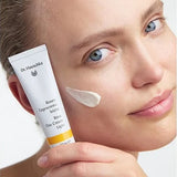 Woman holding a white Dr Hauschka Rose Day Cream Light tube beside her face, with a soft white cream swatch on her cheek.