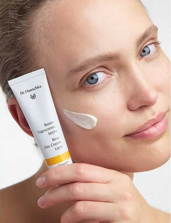 Woman holding a white Dr Hauschka Rose Day Cream Light tube beside her face, with a soft white cream swatch on her cheek.