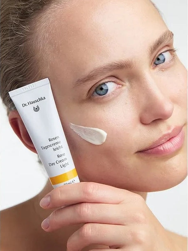 Woman holding a white Dr Hauschka Rose Day Cream Light tube beside her face, with a soft white cream swatch on her cheek.
