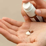 A person dispenses Dr Hauschka Translucent Bronzing Tint 18ml and white cream onto their palm, with a neutral background. Their other hand holds the pump bottle.